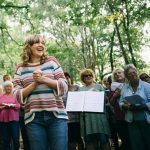 A happy woman with a music stand and a crowd of singers.
